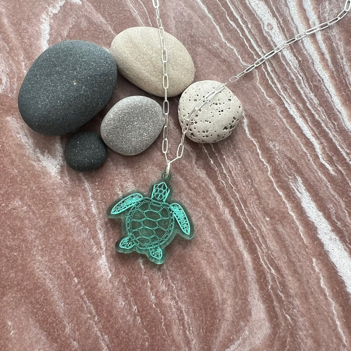 Necklace with a turtle pendant on a marble surface with stones. Sea turtle pendant on a sterling silver chain. Lightweight, Semi-transparent green laser cut acrylic. Lovely to wear to any occasion. 
